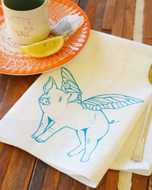beige cloth napkin with a turquoise pig that has wings