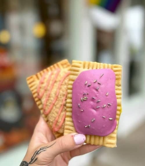 two vegan pop tarts covered in bright purple icing and orange icing drizzle held with one hand on a philadelphia street