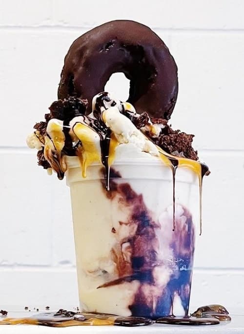large clear cup overflower with vanilla vegan ice cream with swirls of chocolate syrup and topped with a chocolate covered donut