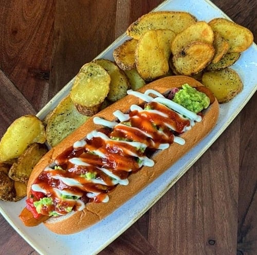 loaded vegan hot dog topped with red hot sauce and white cream sauce next to potatos on a white plate in madrid