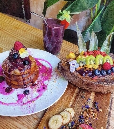 a stack of three vegan pancakes covered in berries and a purple berry sauce next to a purple smoothie and a bowl covered in kiwi, strawberries, and blueberries