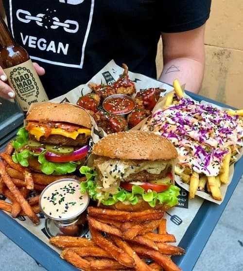 a large tray with two giant vegan burgers topped with lettuce, tomato, and cheese, next to sweet potato fries and a basket of fries covered in  white cream and pickled onions in madrid