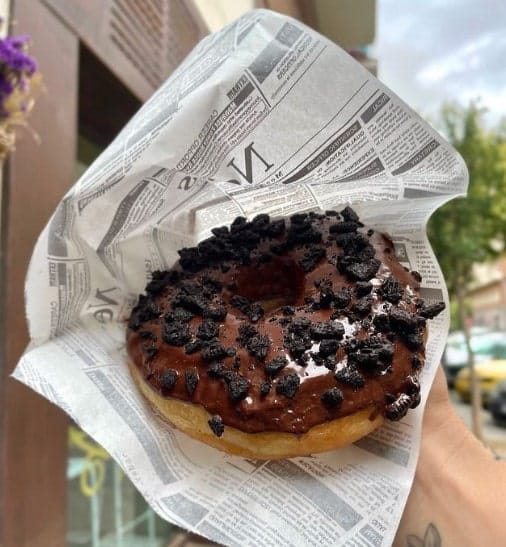 one giant vegan donut topped with chocolate and chocolate cookies held in a newspaper wrapper  in madrid