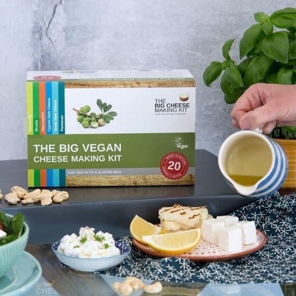 a box containing a vegan cheese making kit in the background in front of vegan cheeses and one hand pouring a vegan liquid cheese
