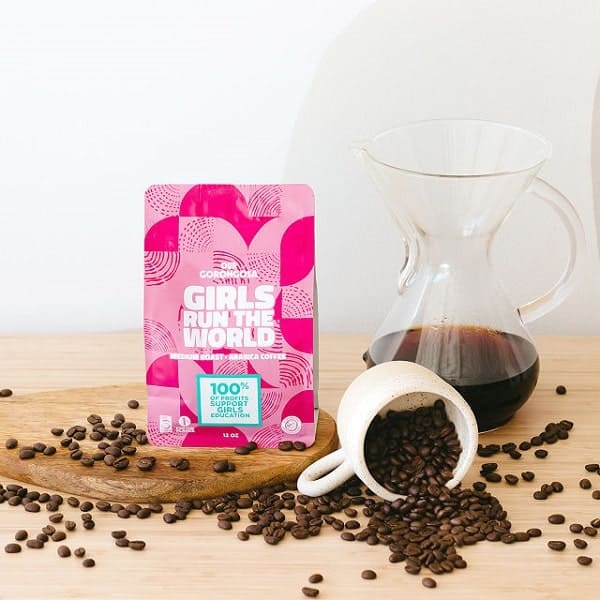 a pink bag of girls run the world coffee beans on a wood table covered in coffee beans next to a coffee mug