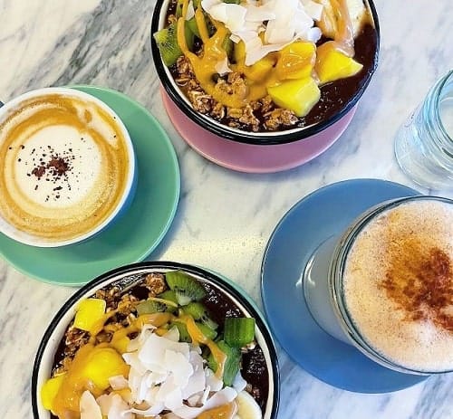two coffees topped with white foam next to two bowls filled with coconut, fruit, and a peanut butter sauce in vienna