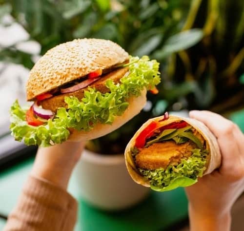 two hands holding a large vegan burger with leafy green lettuce hanging out of the bun and a vegan wrap with a little lettuce and tomato showing in krakow