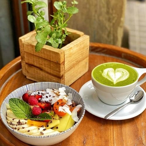 a small table with a bowl of fruit on top of an acai base next to a green matcha drink with a plant behind it at cafe equilibrium in barcelona