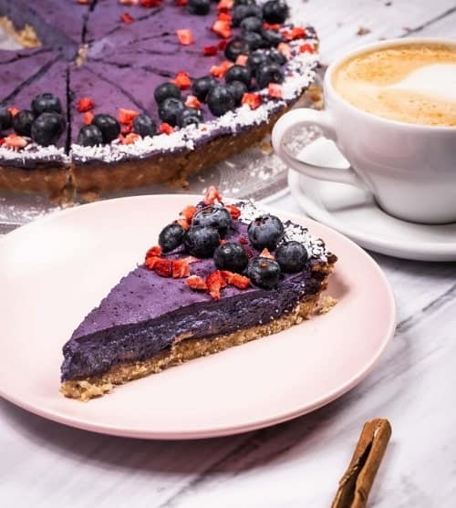 a bright purple slice of vegan blueberry cake on a white plate in front of a larger blueberry cake and next to a cup of coffee in krakow