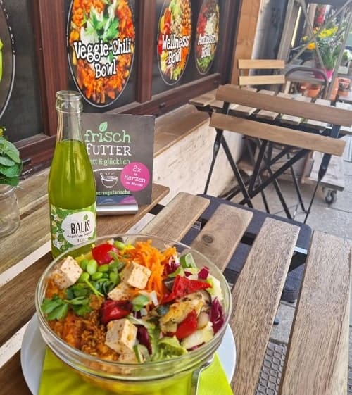 a small bowl filled with tofu, greens, carrots and more on an outside wood table next to a soda bottle in munich