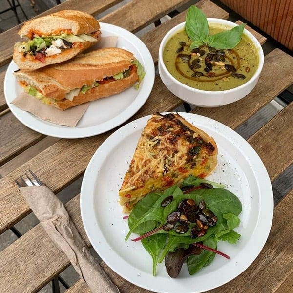 vegan sandwich, quiche, and soup in a triangle pattern on a wooden outdoor table at lucifer lives in brussels