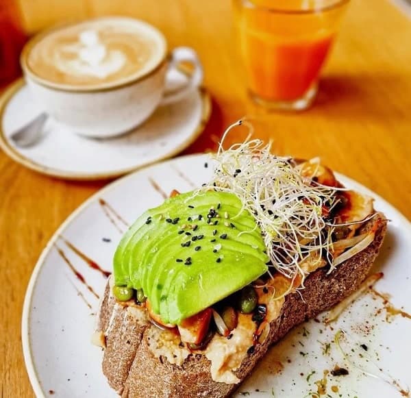 vegan avocado toast next to a cup of coffee and a juice on a light wood table at that's toast in bruges