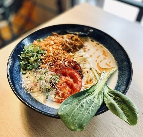 black bowl on a wood table filled with vegan ramen, a light yellow broth, bok choy leaves, and roasted tomato in krakow