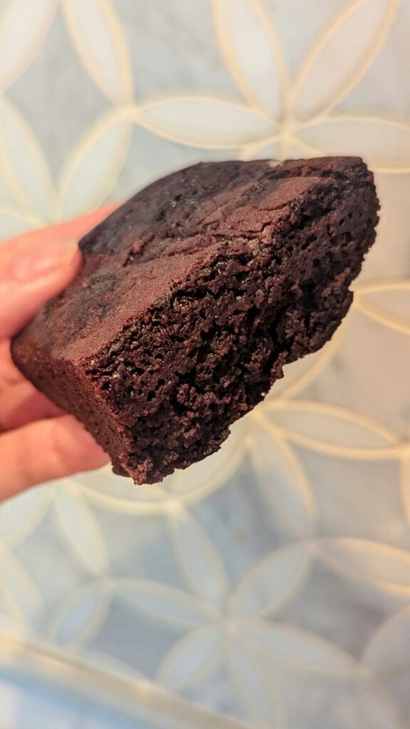 a thick dark chocolate vegan brownie held in front of a marble wall from good cakes and bakes in detroit