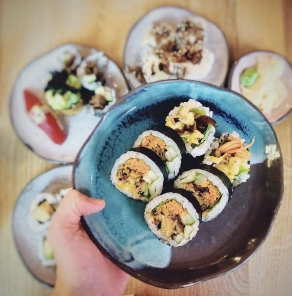a blue and black plate held filled with six vegan sushi rolls held above other japanese fare at youmiko in warsaw