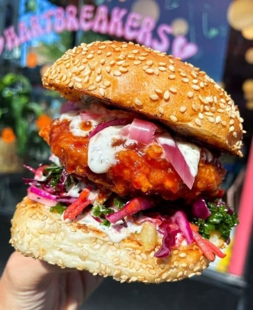 a giant vegan fried chicken sandwich held with one hand covered in bbq sauce and a white cream sauce in nyc