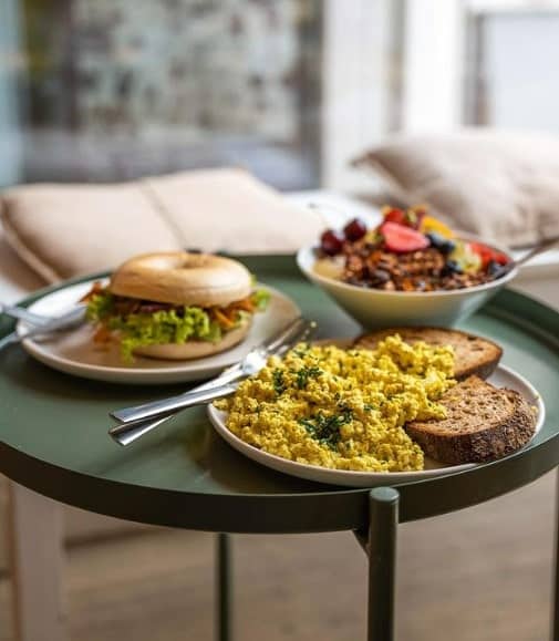 vegan breakfast spread with a plate of scrambled eggs and toast, bagel breakfast sandwich, and a granola and fruit bowl at plant base in berlin