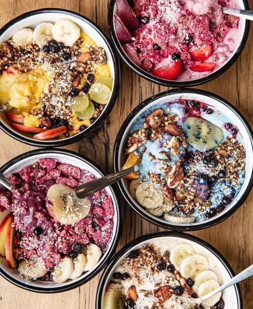 five different colorful bowls filled with fresh fruit, granola, oatmeal, and seeds on a wood table at wild and the moon in amsterdam