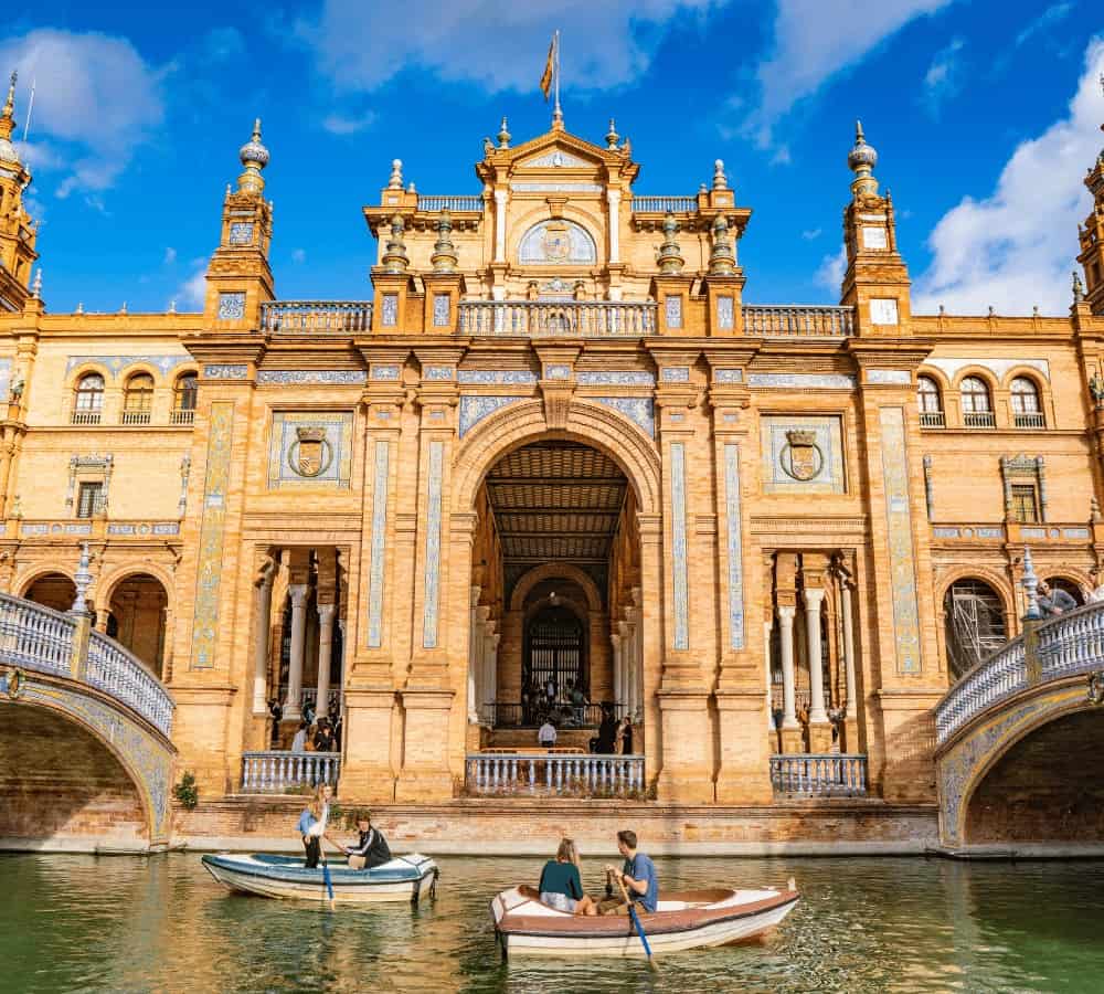 two people rowing boats in the small river that runs through the plaza de espana on a bright day in seville