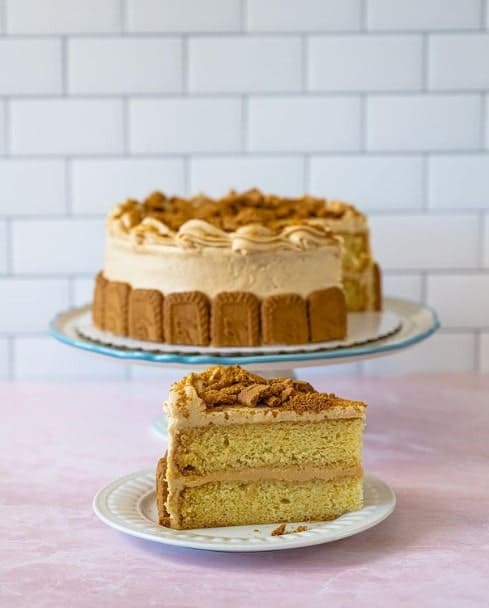 large vegan cake with biscoff cookies lining the edge. one large cake is in the backgound with a slice in front of it