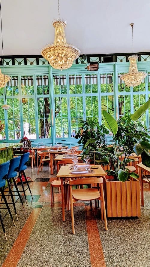 inside of bastion park cafe with two hang crystal chandeliers, light wood tables and chairs and a turquoise wall of windows