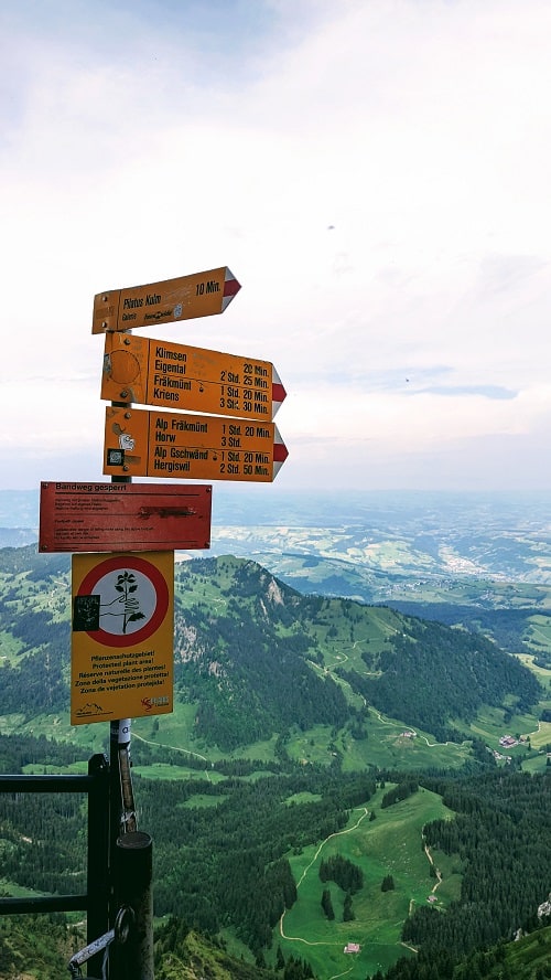 orange hiking trail sign on mount pilatus with a cloudy background of green meadows and lucerne