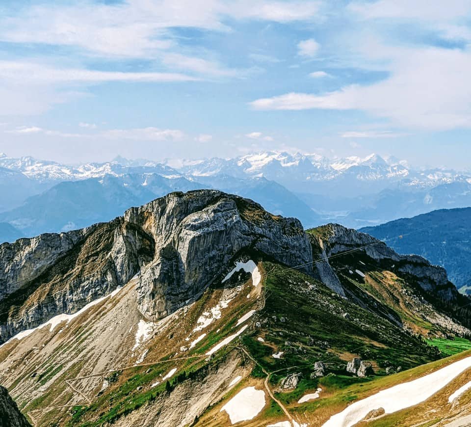 partly cloudy view from mount pilatus that shows green and snow covered mountain and the alps in the background