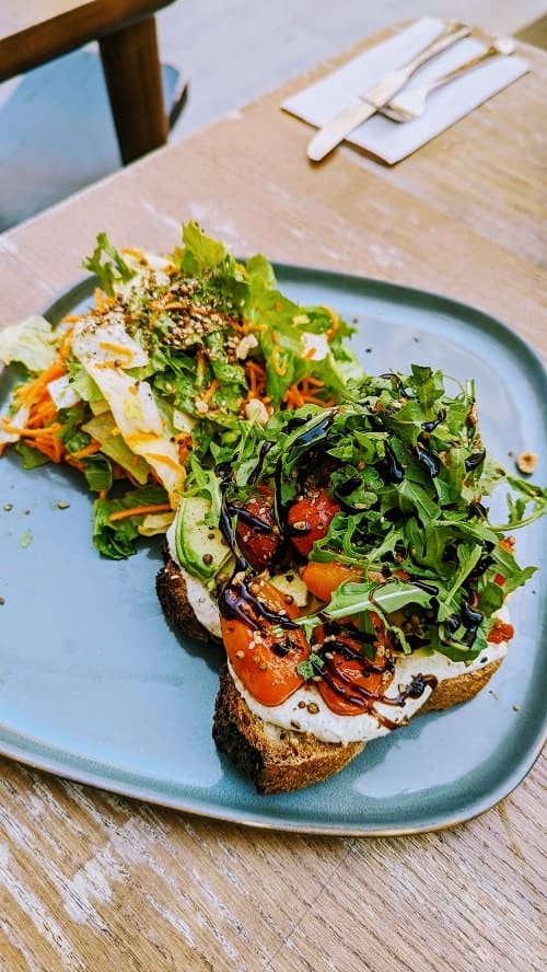vegan and glutenfree toast covered in leafy arugula, vegan cashew cheese, roasted tomatoes and a balsamic drizzle on a blue square plate from ou bien encore in geneva