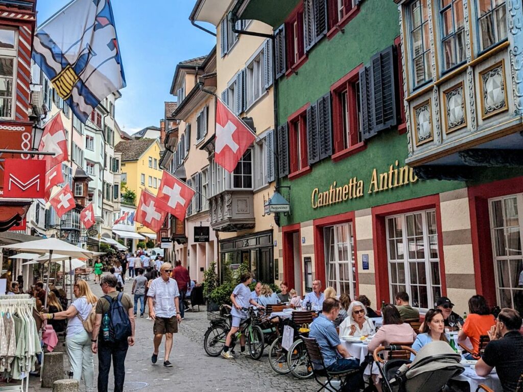 a narrow street in zurich's old town filled with people eating at small tables and shopping with red and white swiss flags overhead