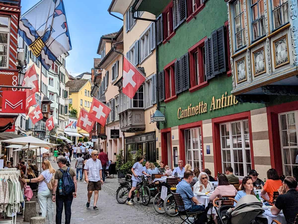 a narrow street in zurich's old town filled with people eating at small tables and shopping with red and white swiss flags overhead