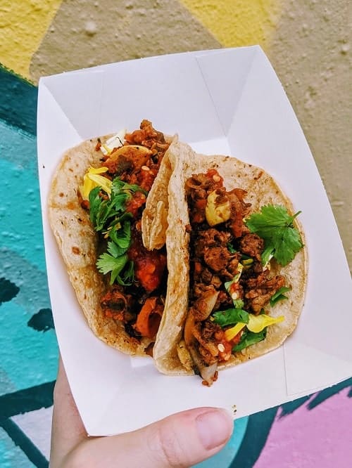 two vegan tacos held in a small white tray in front of a colorful wall mural in detroit