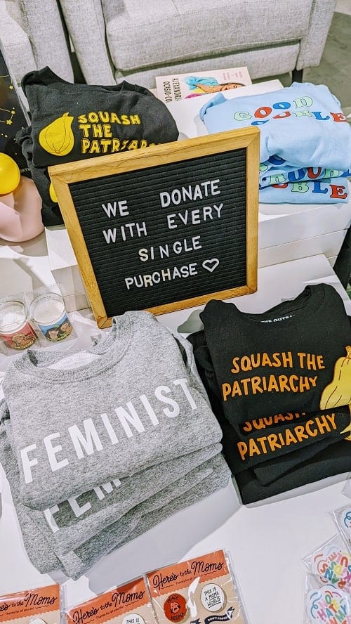 light gray feminist sweatshirts and black squash the patriarchy tees folded in piles on a white table at the outrage in dc