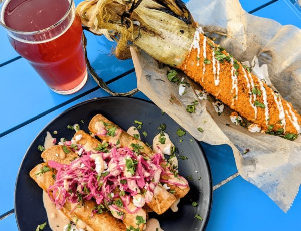 vegan taquitos covered in pickled onions on a black plate next to a basket with mexican street corn and a glass of pink berry kombucha
