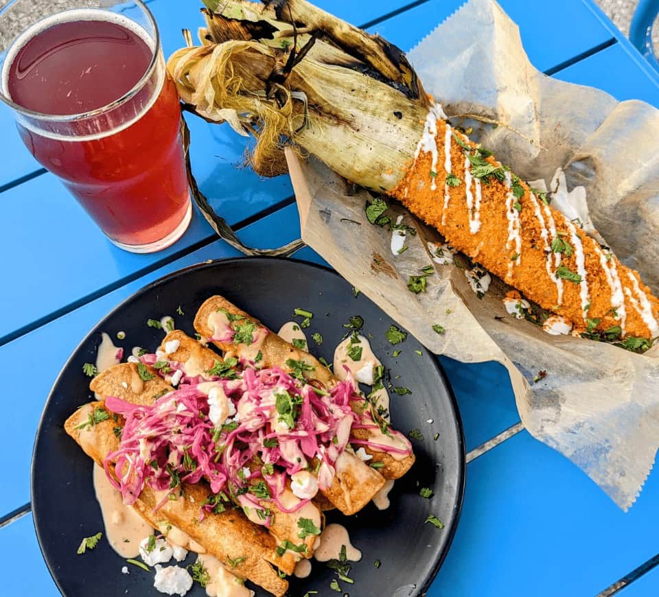 vegan taquitos covered in pickled onions on a black plate next to a basket with mexican street corn and a glass of pink berry kombucha