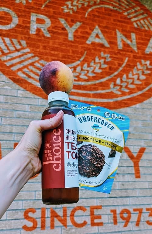 michigan peach on top of a bottle of choice cherry tea and a bag of vegan chocolates in front of the orange oryana co-op sign in traverse city