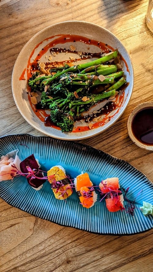 bowl of spicy broccoli stems covered in a chili sauce and almonds and in front if it vegan sushi nigiri on a blue leaf plate at kusaki in brighton