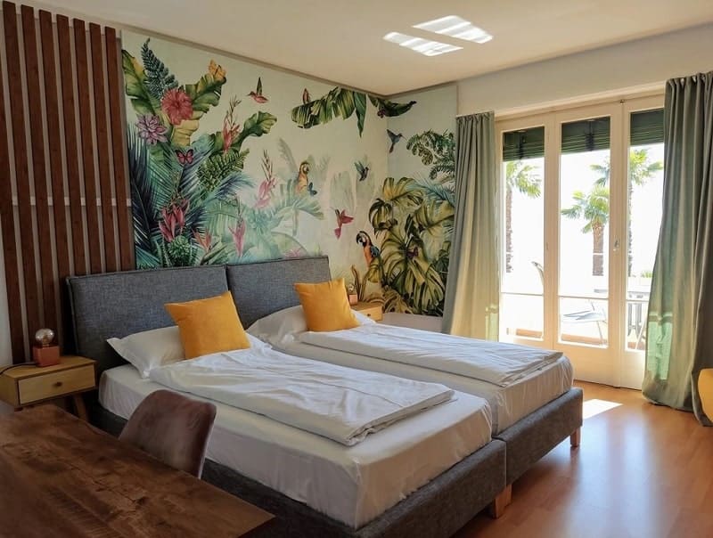 large guestroom with a double bed, a floral jungle mural behind the bed and a large sunny window at the hotel garden primavera in switzerland