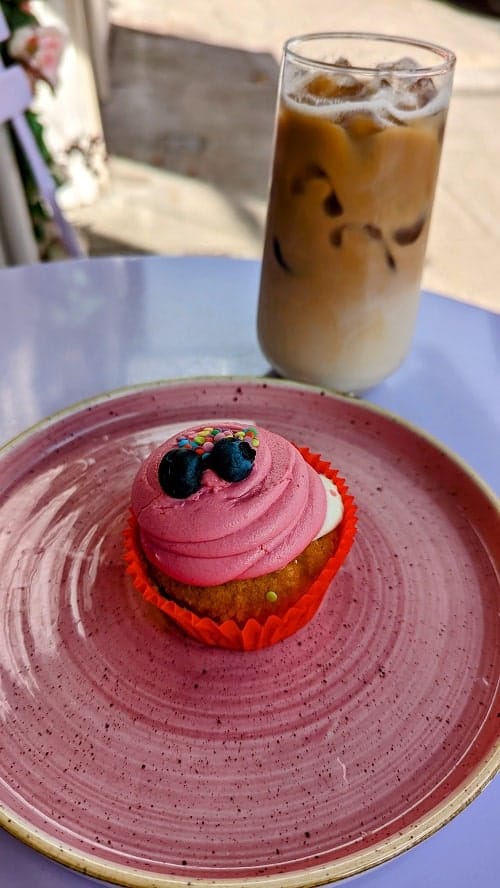 vegan gluten free cupcake covered in pink buttercream and topped with two blueberries sitting on a pink plate with an iced coffee behind it at plant blonde glasgow