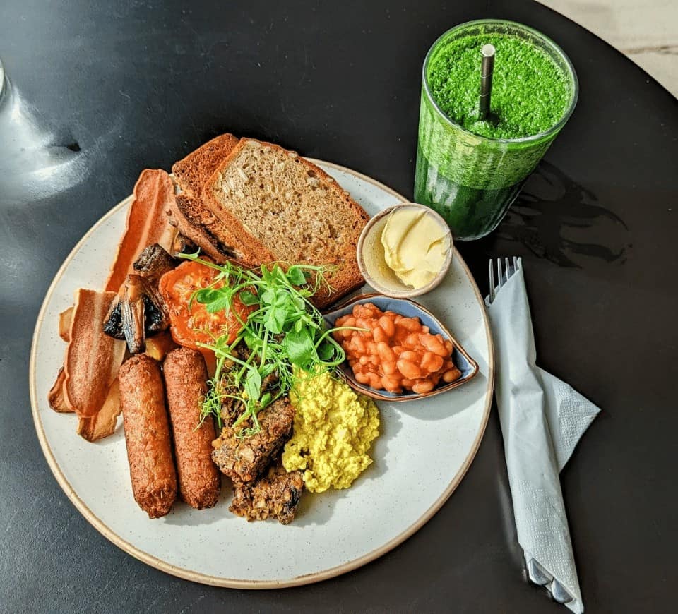 veagn scottish breakfast with sausages, toast, beans, tomatoes on a white plate next to a green smoothie at holy cow in edinburgh