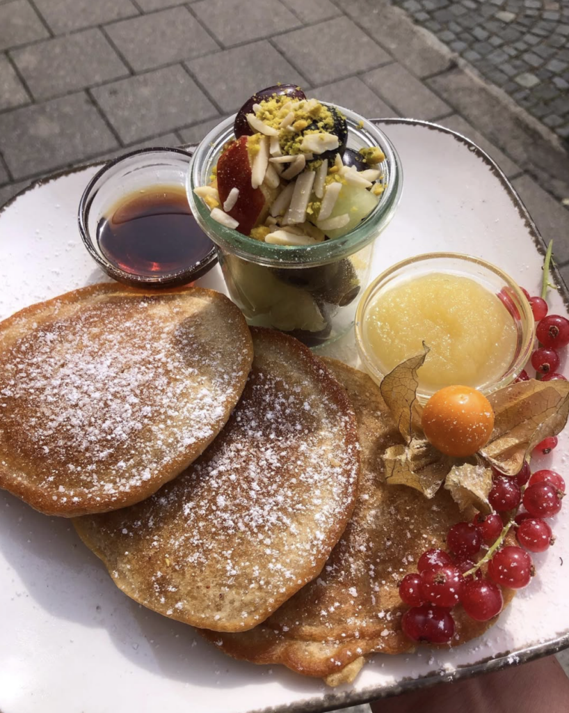 Vegan breakfast at Deli Kitchen Munich featuring pancakes with powdered sugar, fresh fruit, applesauce, and maple syrup on a plate outdoors.