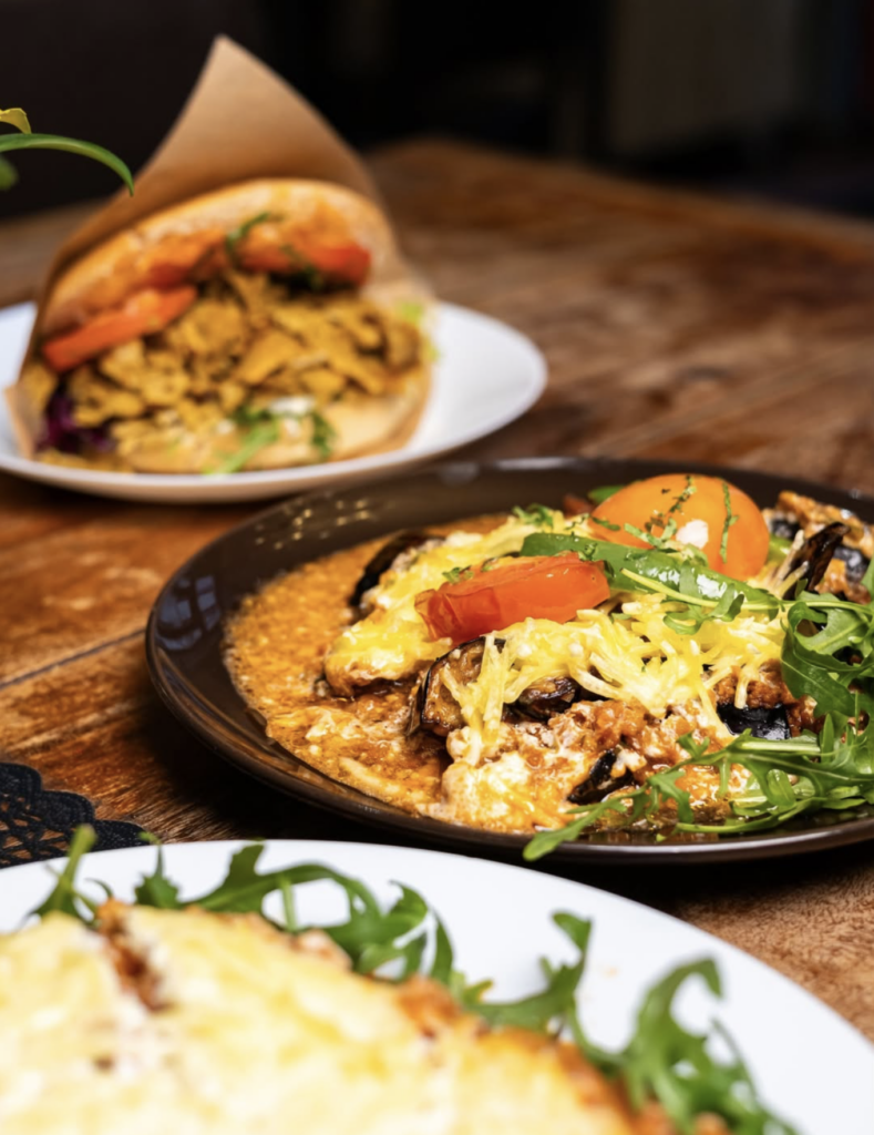 Vegan Middle Eastern dishes at Erbil’s Restaurant featuring eggplant with tomato and herbs, served alongside a stuffed sandwich on a wooden table.