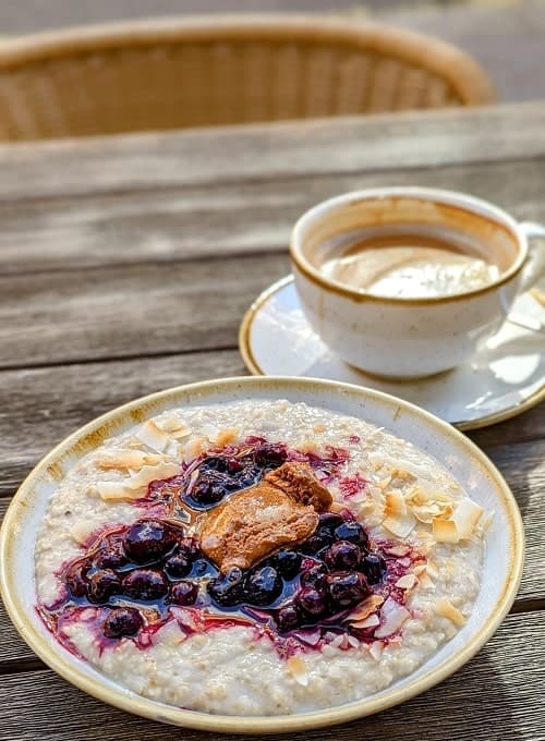 a large white bowl filled with creamy oat porridge and topped with blue berries and alond butter in front of a cup of coffee in london