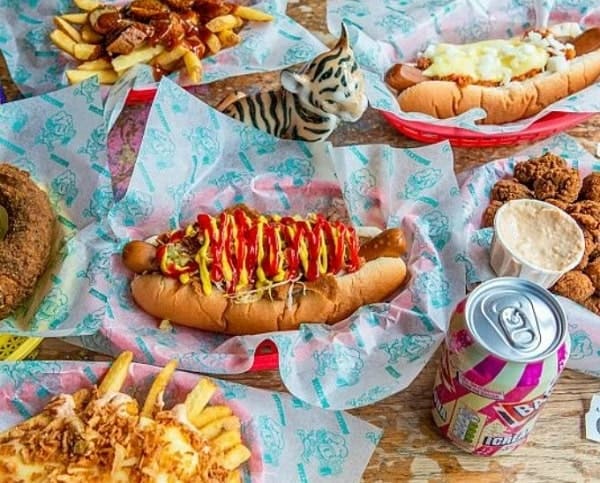five baskets filled with vegan hot dogs covered in ketchup, cheese, onions along with loaded french fries covered in cheese next to a can of soda in brighton