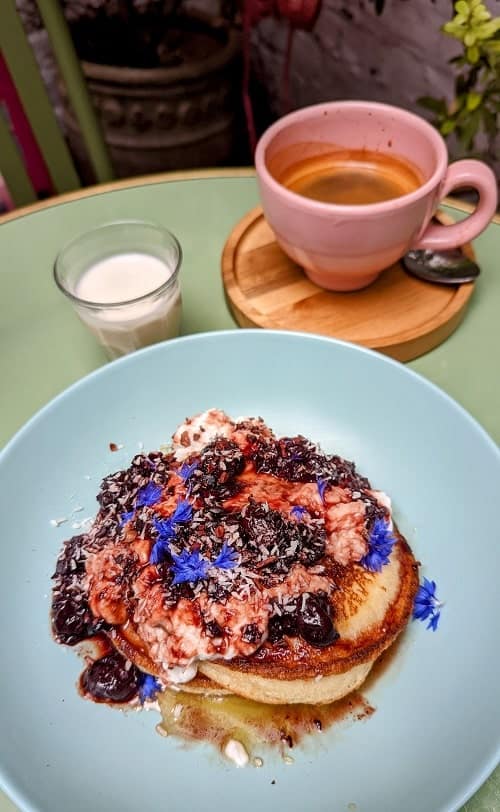 a large light blue plate with a small stack of golden vegan pancaked topped with berry compote, white coconut yogurt and edible flowers in front of a pink mug with coffee in london