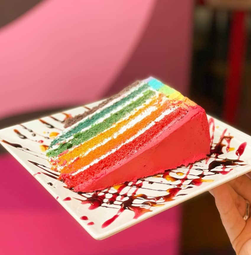 a slice of colorful vegan rainbow cake on a white plate covered in chocolate and strawberry sauce in madrid