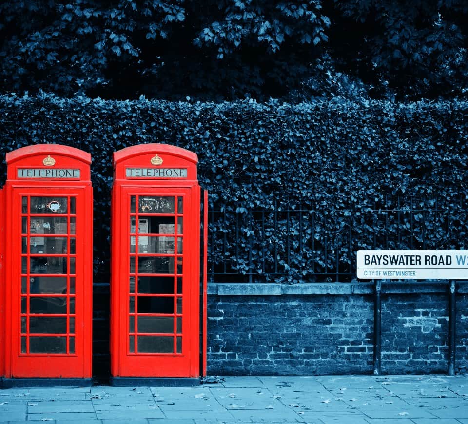 two bright red london telephone booths next to each other in front of a dark hedge on baystwater street