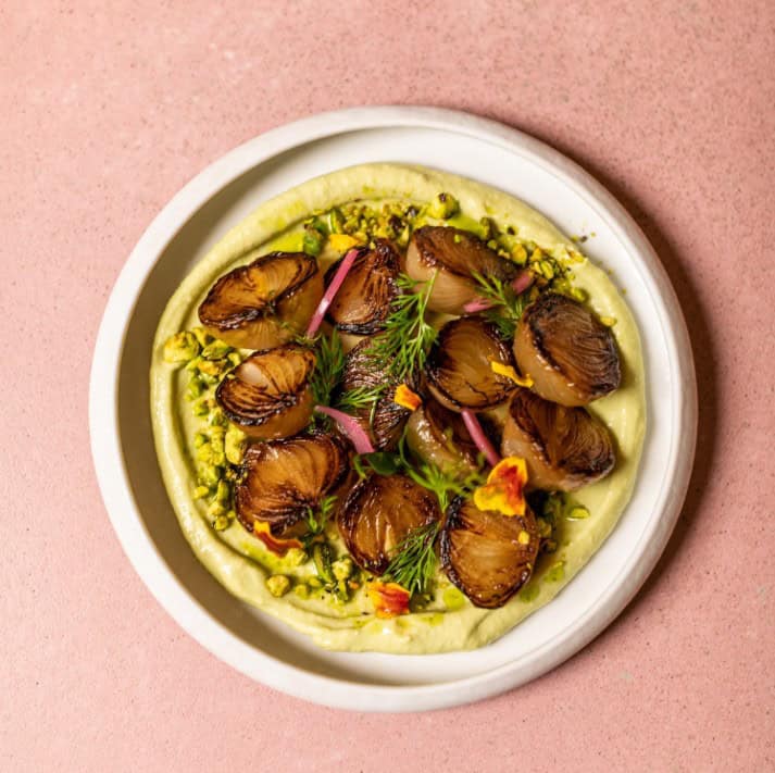Miso Butter Onions are miso-glazed French onions, accompanied by a smooth avocado cream, cilantro oil, roasted pistachios, and pea shoots. at the vegan restaurant mudra in madrid