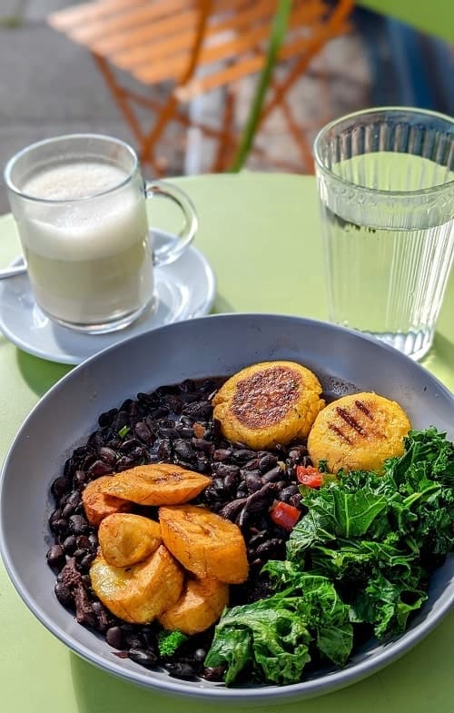 a large gray bowl filled with bright greens, plantains, black beans and corn cakes in front of a water glass and a tea cup in london