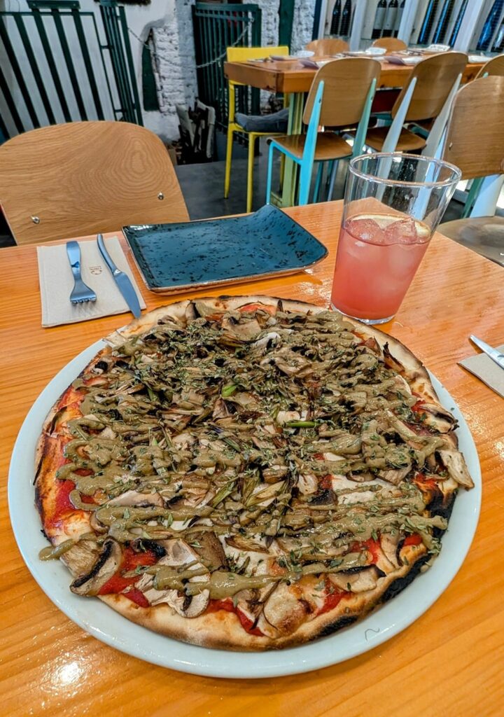 large vegan and gluten free pizza topped with greens and pistachio cream next to a light pink drink at pizzi and dixie in madrid