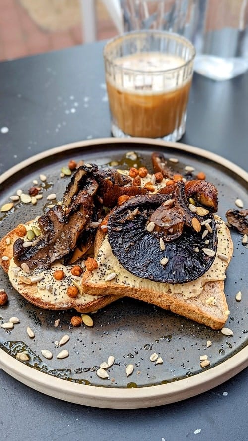 three slices of vegan and gluten free toast on a black plate topped with a mix of cooked mushrooms and hummus with a small coffee in the background in brighton